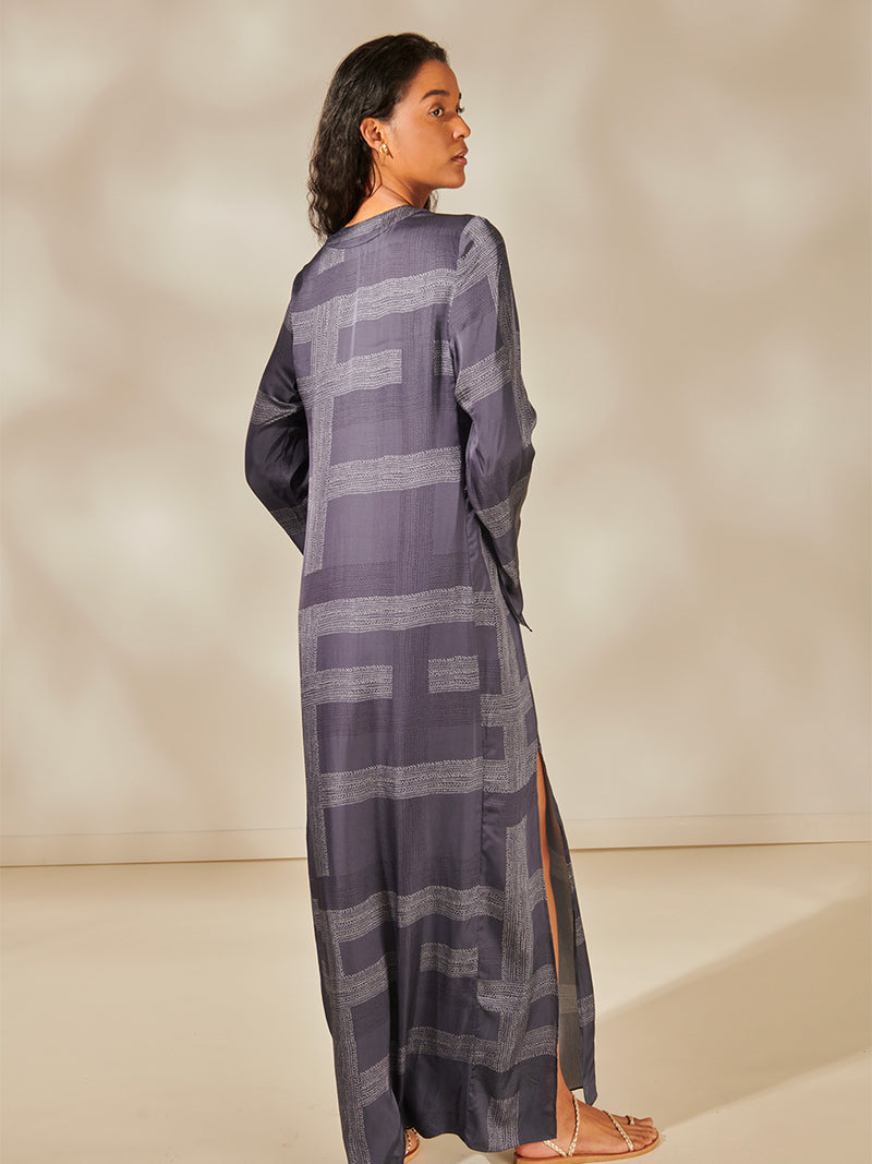 Back view of a Woman Standing Wearing Theodora Column Dress Featuring an elegant hand-sketched chevron pattern, featuring alternating neutral and charcoal stripes set against a cool steel blue background.