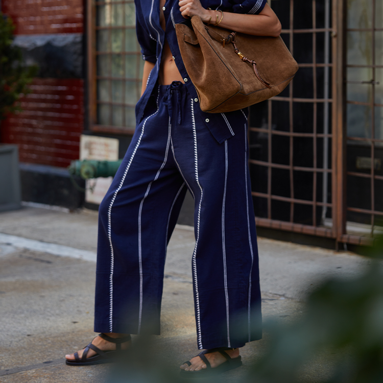 Close on woman's legs wearing lemlem handwoven pants and matching button down shirt in blue with graphic white stripes.