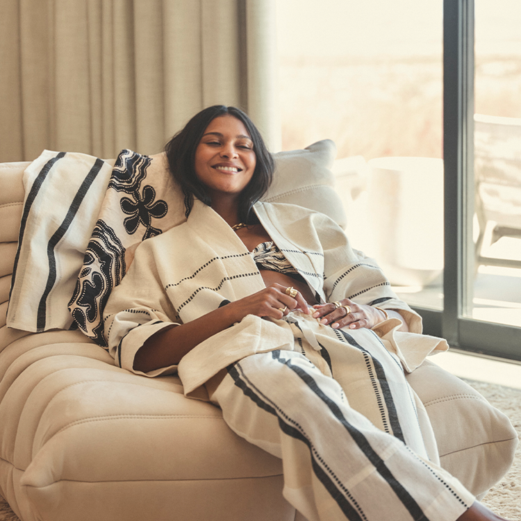 Woman sitting on a couch wearing lemlem handwoven jacket and pants, smiling.