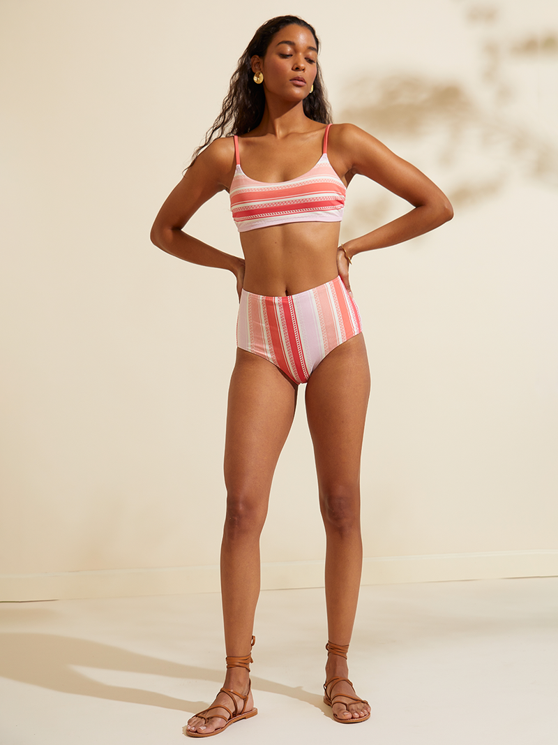 Woman standing wearing the Eshal High Waist Bikini Bottom featuring white doted stripes with gradiant orange and tangerine bands on a lilac and white background.