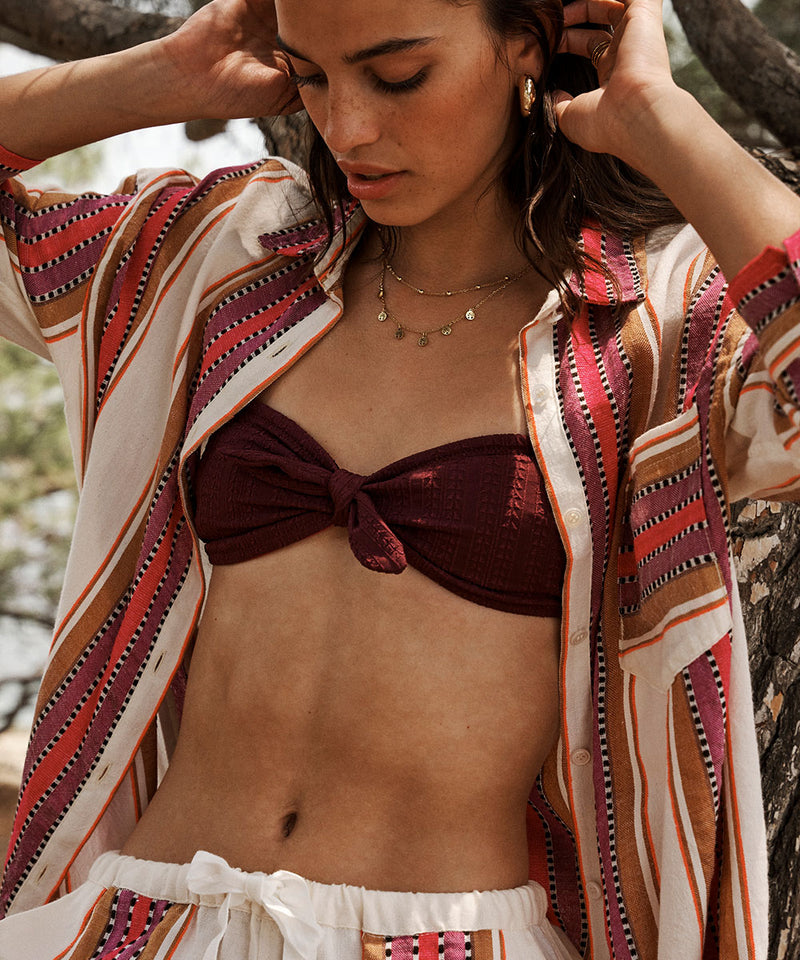 Woman standing against a tree wearing a burgundy bandeau bikini top and an open shirt in cream with brown and burgundy stripes.