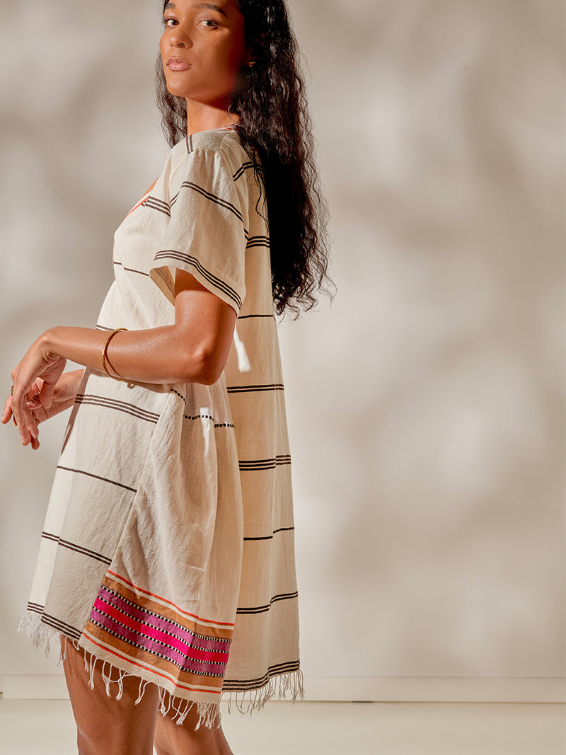 Side View of a Woman Standing Wearing Amina V Neck Caftan featuring stripe pattern in magenta, ochre, and berry tones, delineated by black and white dots and accented with a splash of neon orange on natural cotton background
