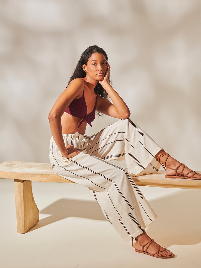 Woman sitting on the bench Wearing Desta Wide Leg Pants featuring stripe pattern in magenta, ochre, and berry tones, delineated by black and white dots and accented with a splash of neon orange on natural cotton background and Jordanos Burgundy Front Tie Bikini Top