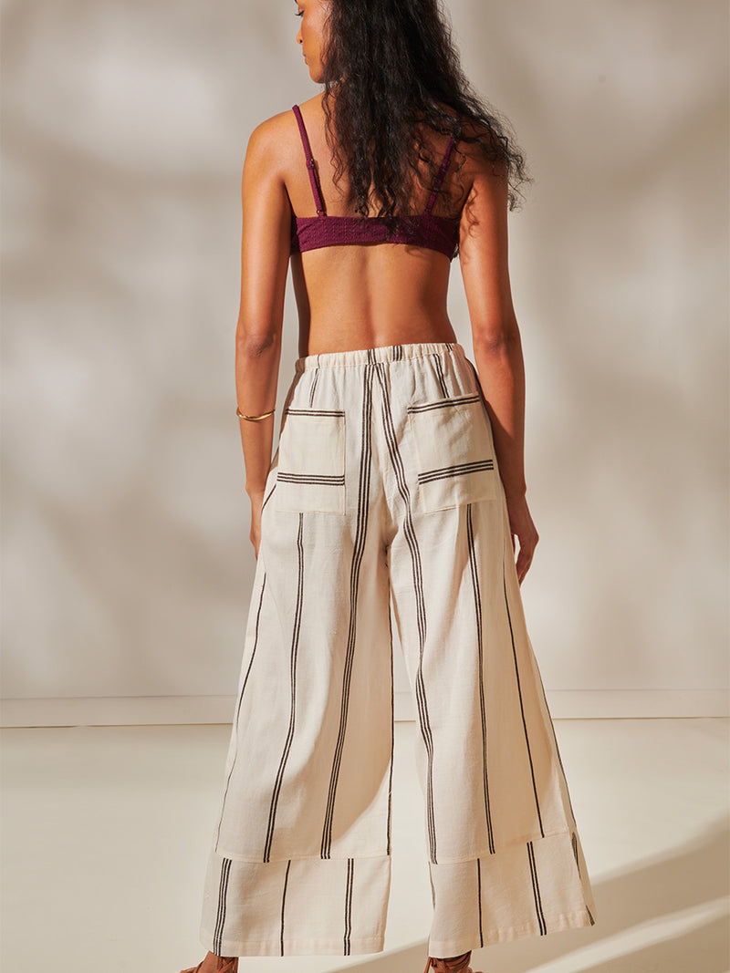 Back View of a Woman standing Wearing Desta Wide Leg Pants featuring stripe pattern in magenta, ochre, and berry tones, delineated by black and white dots and accented with a splash of neon orange on natural cotton background and Jordanos Burgundy Front Tie Bikini Top