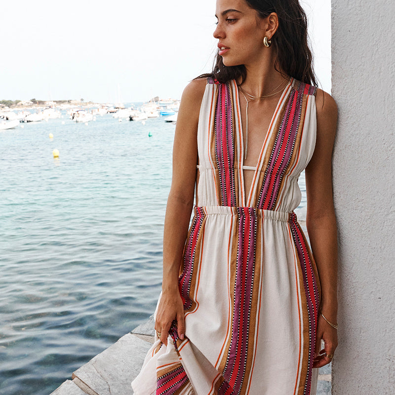 Woman standing against a white wall with the sea in the background, wearing a long plunge neck dress in cream with brown and burgundy stripes