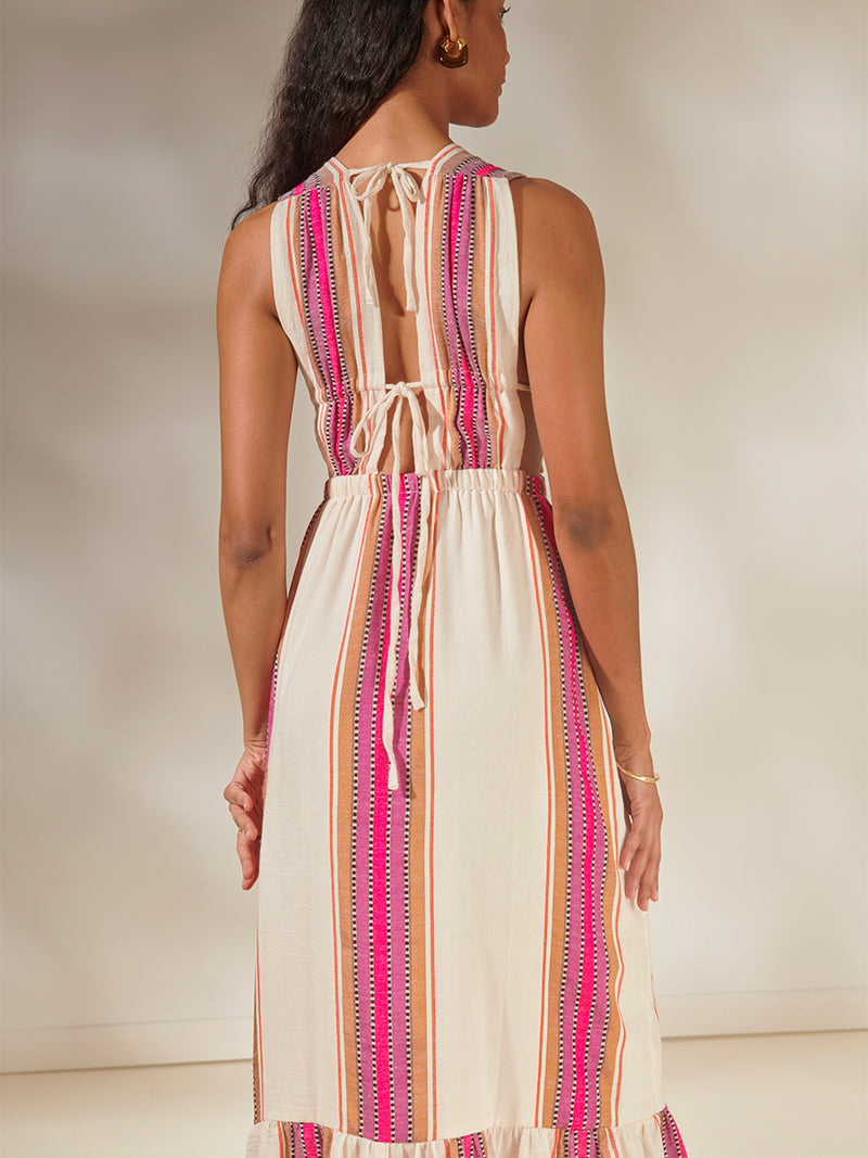 Back View of a Woman Standing Wearing Lelisa V Neck Dress featuring stripe pattern in magenta, ochre, and berry tones, delineated by black and white dots and accented with a splash of neon orange on natural cotton background