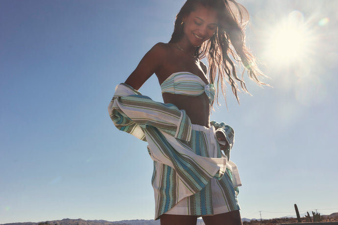 Woman in outdoors with mountains in the background wearing lemlem handwoven shirt and shorts featuring striped pattern in green, mint and sage colors