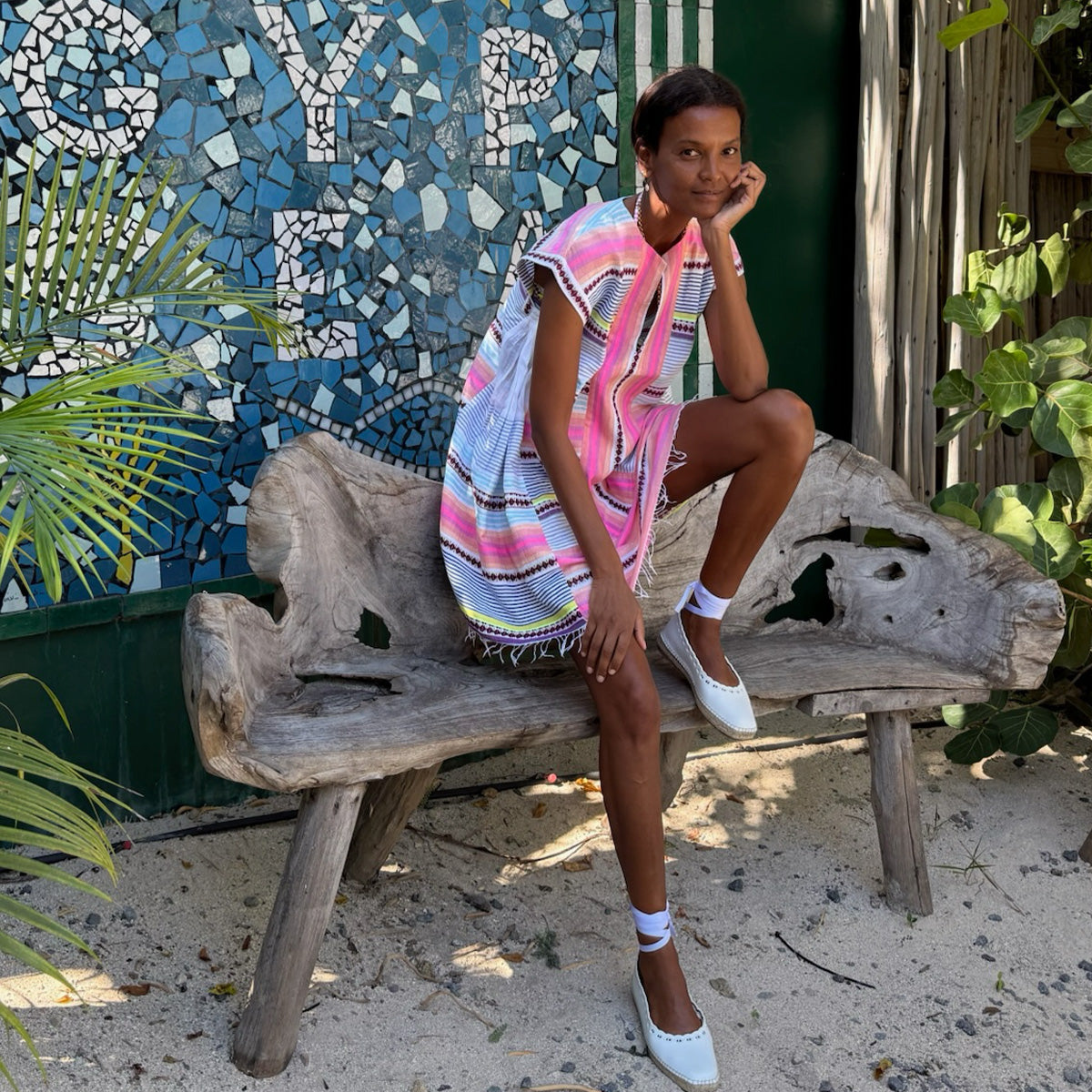 Liya Kebede wearing lemlem handwoven ELINA Caftan featuring tibeb and striped pattern sitting on a wooden bench with a mosaic wall and plants in the background