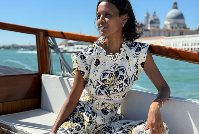 Liya Kebebe sitting on a boat deck in Venice wearing a lemlem linen crop top and matching linen high waist pants in white with lue and yellow tile prints. 