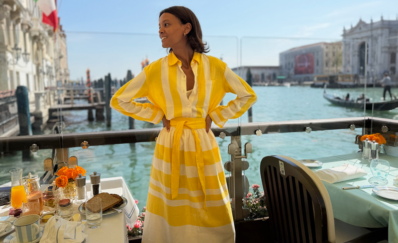 Liya Kebebe in a yellow lemlem handwoven shirt and matching skirts standing by a waterfront with tables set for a meal.