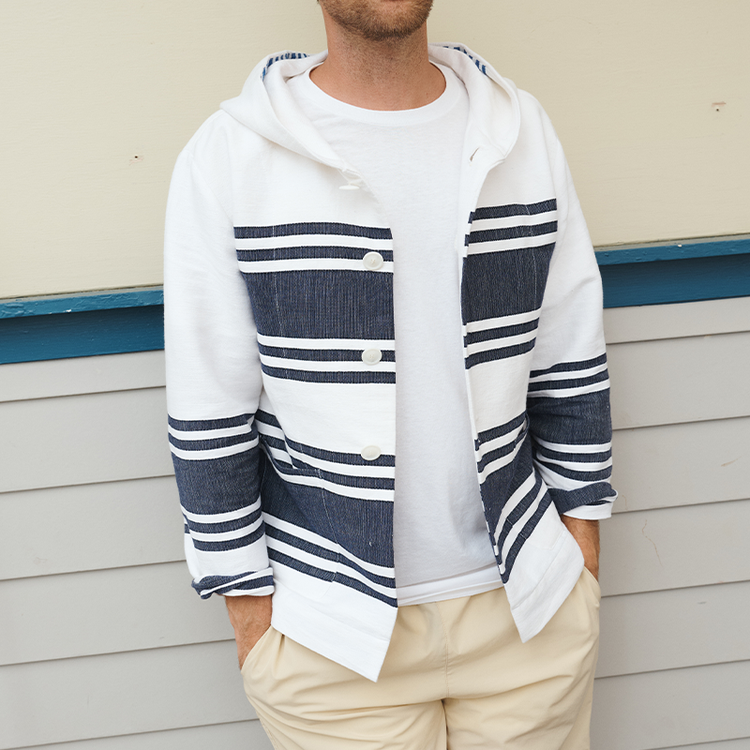 A man wearing a white hooded cardigan with navy blue horizontal stripes, layered over a white t-shirt and beige pants, standing against a beige and white paneled wall.