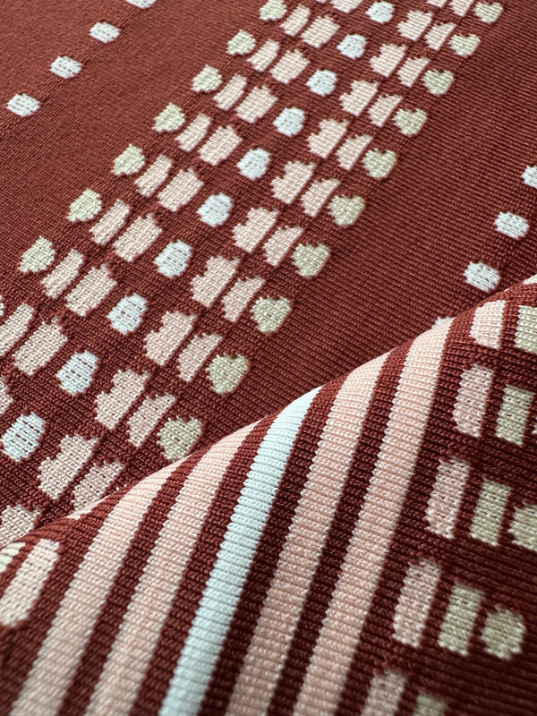 Close-up of a woven textile with a rust red background, cream and pale blue geometric motifs, and diagonal striped sections in cream, blush pink, and pale blue.