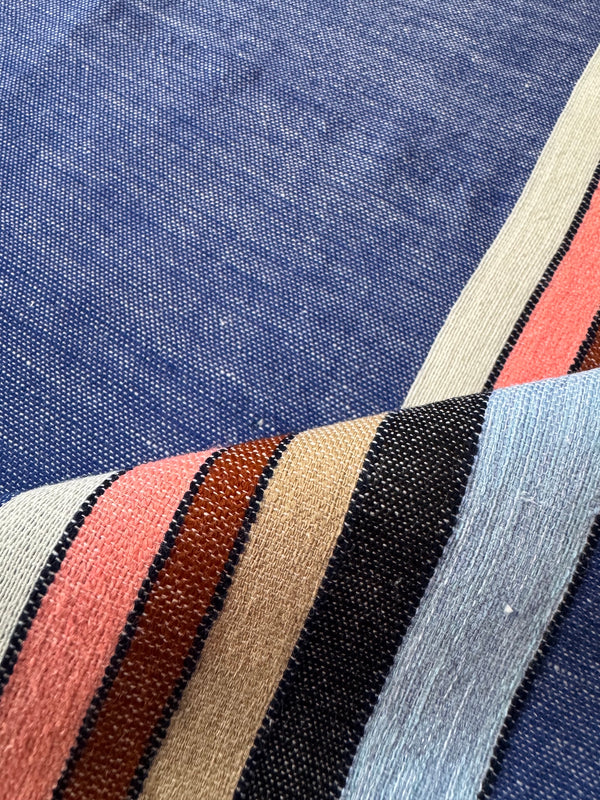 A close-up of lemlem handwoven textile with a dominant medium blue field and bold diagonal stripes in cream, coral pink, rust brown, black, and pale blue.