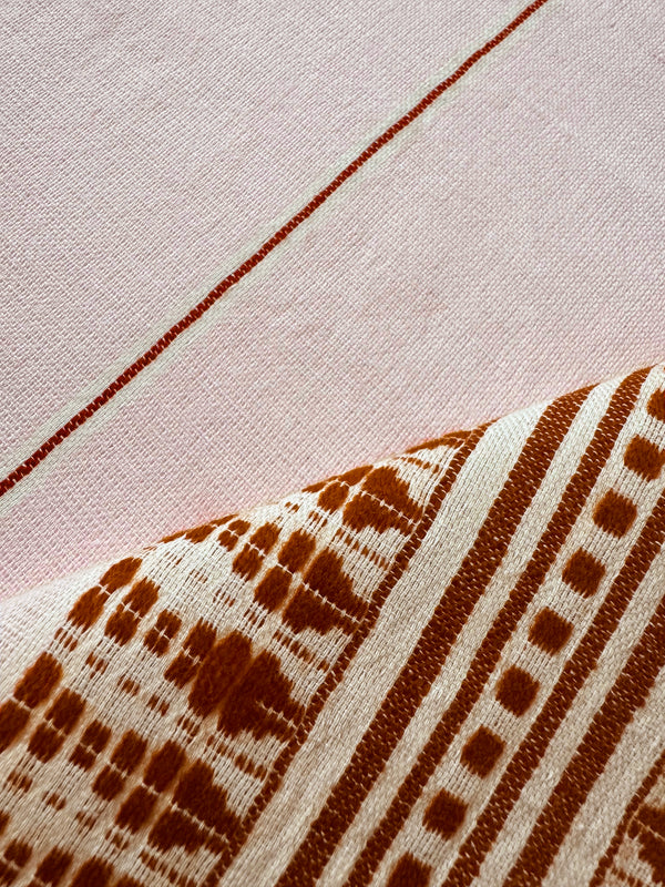 Close-up of a handwoven textile with a pale pink background and a folded section showing a rust-brown geometric pattern on an off-white base.