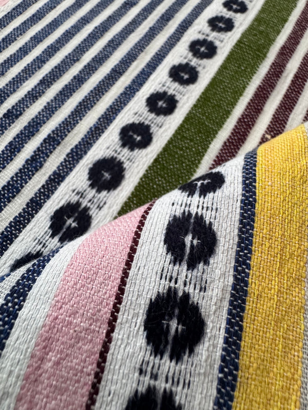 Close-up of lemlem handwoven textile with vertical stripes in navy, white, olive green, burgundy, pink, and mustard yellow, overlaid with a patterned section featuring black ikat-like motifs on a white base.