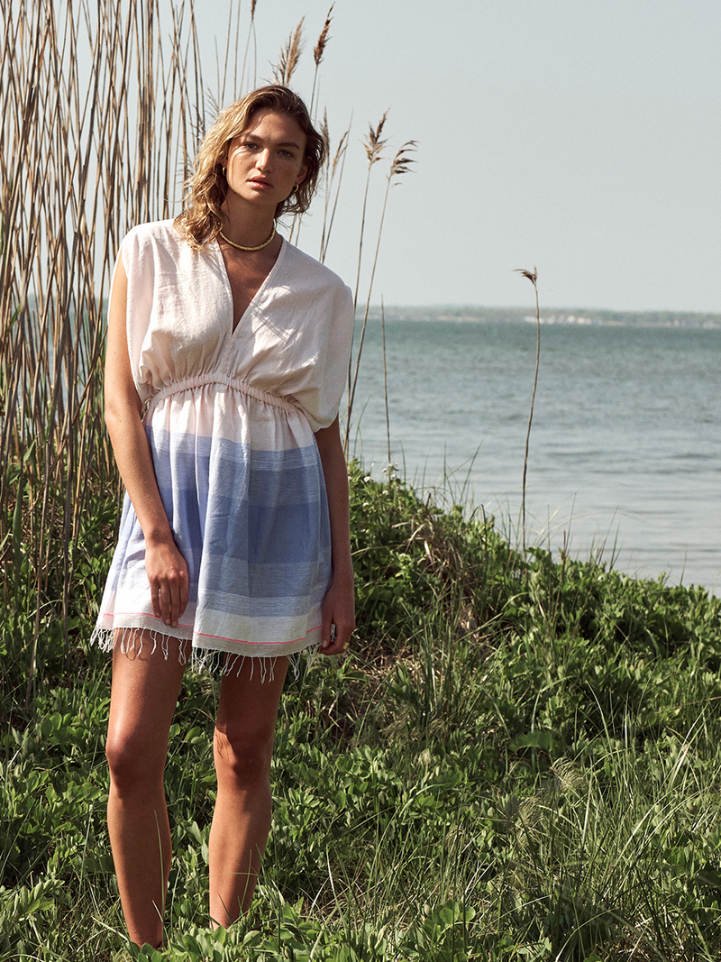 Woman Standing on the grass on the Fire Island Beach Wearing Jelba Short Plunge Neck Dress featuring gradient color block design of nine shades of soft pink and blues highlighted by a bright pink mini stripe at the edge of the dress.
