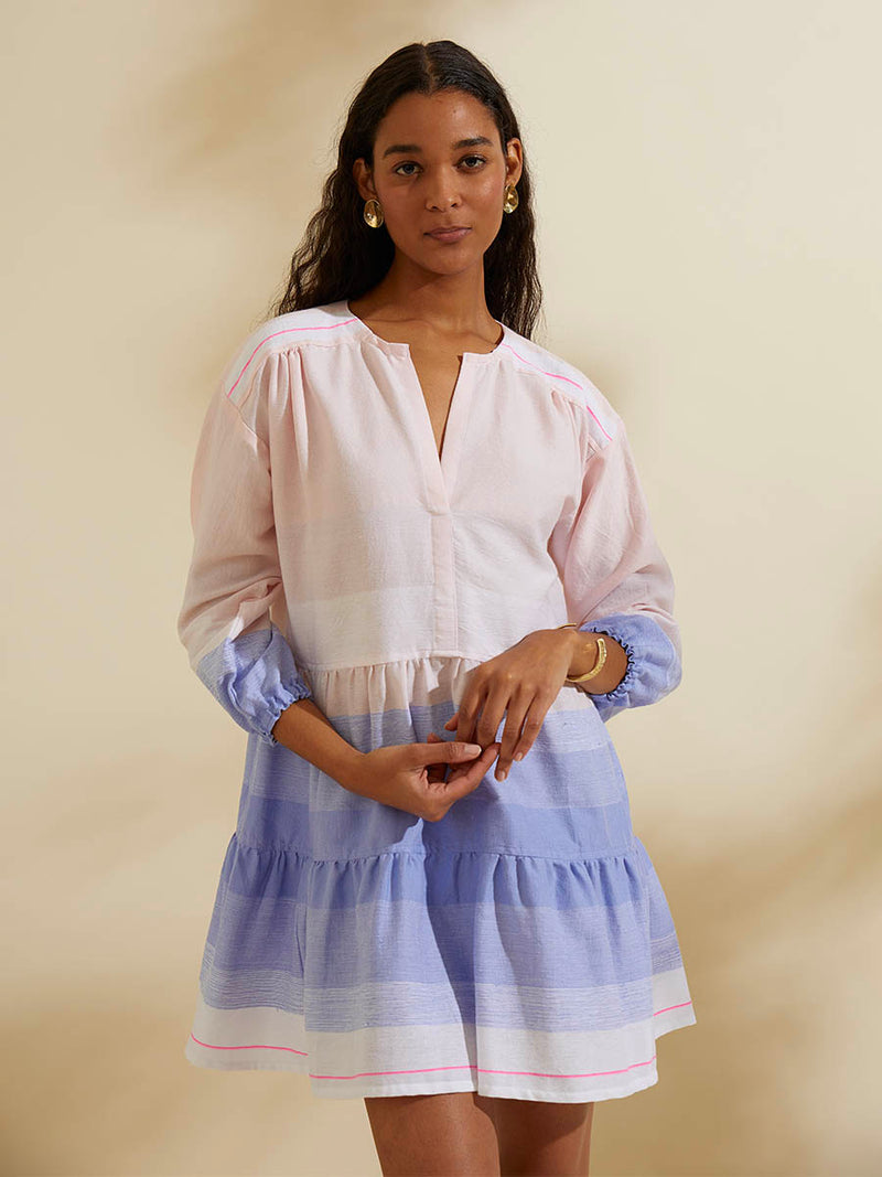Woman standing wearing the Jelba Popover Dress featuring gradient color block design of nine shades of soft pink and blues highlighted by a bright pink mini stripe at the edge of the dress.