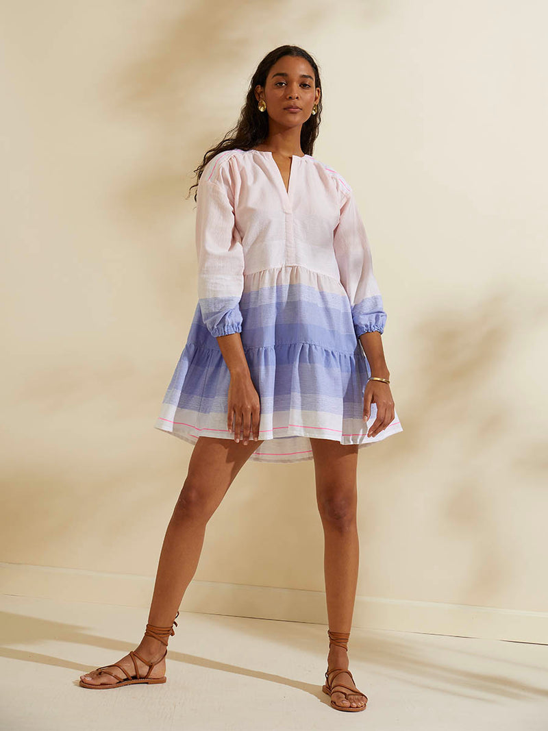 Woman standing wearing the Jelba Popover Dress featuring gradient color block design of nine shades of soft pink and blues highlighted by a bright pink mini stripe at the edge of the dress.
