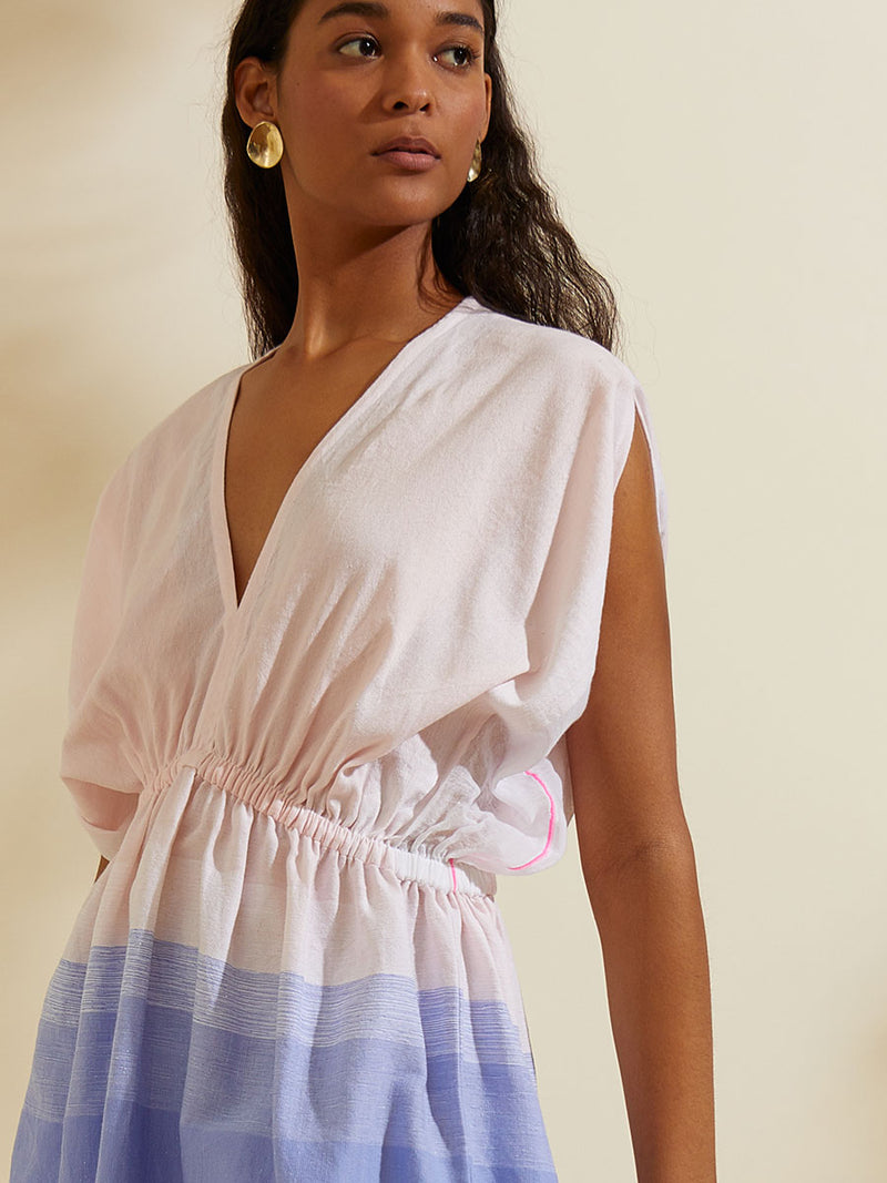 Close up shot of a Woman standing  wearing the Jelba Short Plunge Neck Dress featuring gradient color block design of nine shades of soft pink and blues highlighted by a bright pink mini stripe at the edge of the dress.