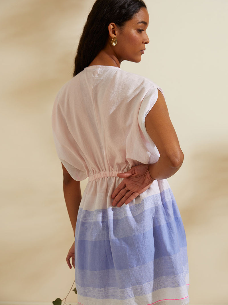 Back view of a woman standing wearing the Jelba Short Plunge Neck Dress featuring gradient color block design of nine shades of soft pink and blues highlighted by a bright pink mini stripe at the edge of the dress.