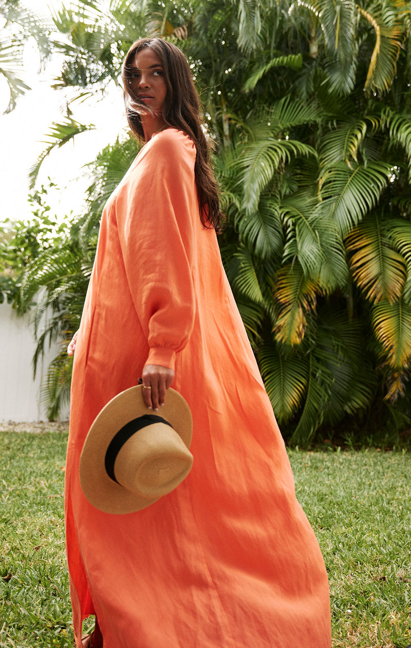  Woman walking and holding a straw hat in her hand wearing the lemlem makeda long dress in coral linen tencel. 