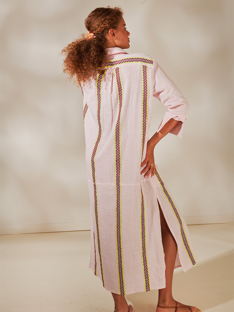 Back View of a Woman Standing Wearing Anata Shirt Dress featuring delicate pink stripes with a bold chevron patterned ribbon, along with muted hues of pink, burgundy, and a bright citrus-orange hue.