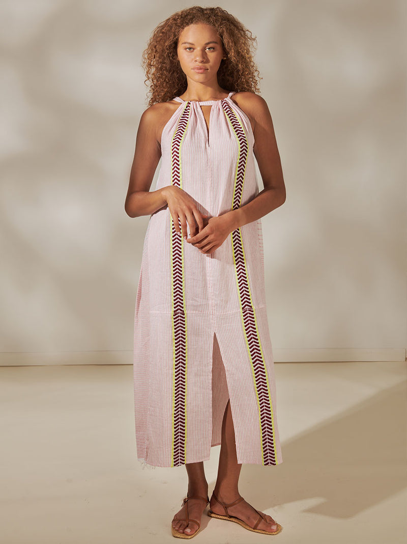 Woman Standing Wearing Ayana Halter Dress featuring delicate pink stripes with a bold chevron patterned ribbon, along with muted hues of pink, burgundy, and a bright citrus-orange hue.