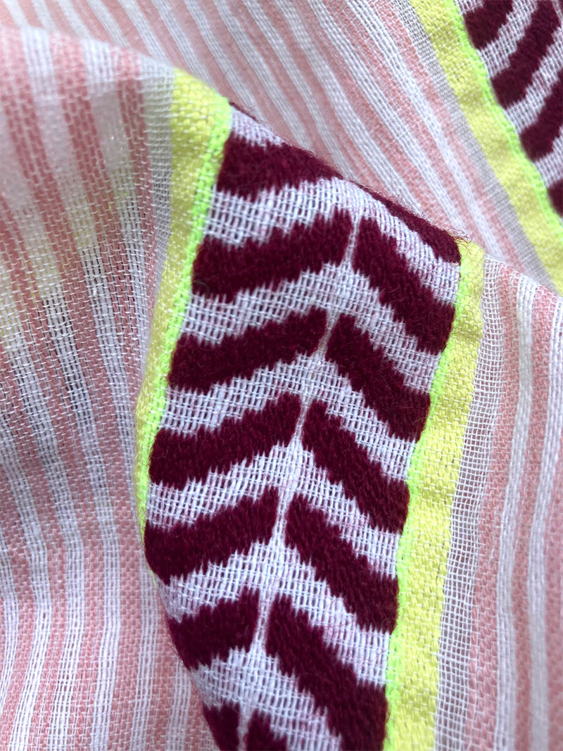 Close up on the fabric of the Edna V Neck Dress featuring delicate pink stripes with a bold chevron patterned ribbon, along with muted hues of pink, burgundy, and a bright citrus-orange hue.