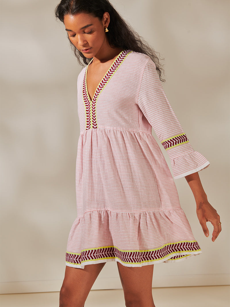 Side View of a Woman Standing Wearing Hanna Flutter Dress featuring delicate pink stripes with a bold chevron patterned ribbon, along with muted hues of pink, burgundy, and a bright citrus-orange hue.