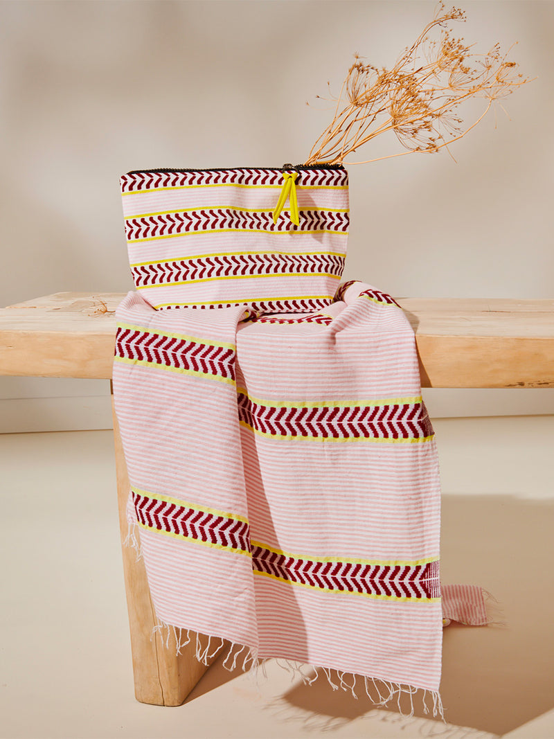 Kesiti Pink Cora Pouch and Kesiti Pink Lema Sarong featuring delicate pink stripes with a bold chevron patterned ribbon, along with muted hues of pink, burgundy, and a bright citrus hue laying on the wooden bench