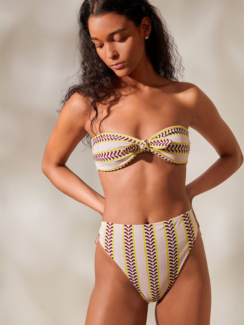 Woman Standing Wearing AVA Bandeau Top and High Leg Bikini Bottom featuring delicate pink stripes with a bold chevron patterned ribbon, along with muted hues of pink, burgundy, and a bright citrus hue.