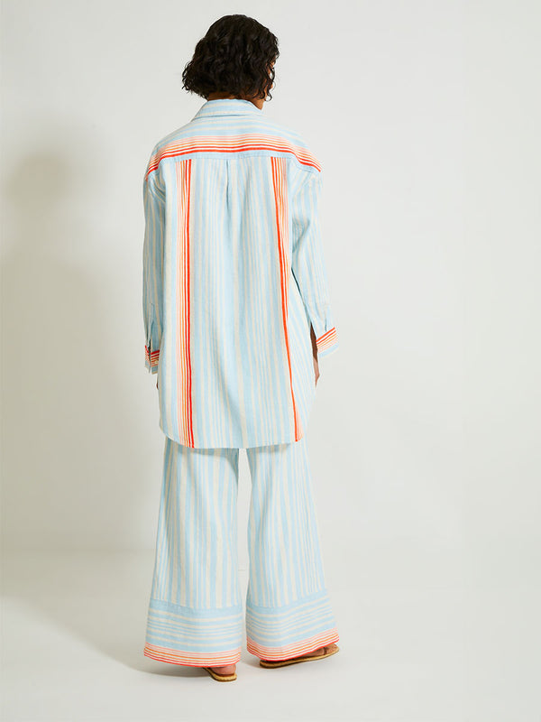 Back of a Woman Standing Wearing lemlem Mariam Shirt Featuring soft, variegated two-color stripe in sky blue and cream, complemented by placed bands of bright orange and tangerine.