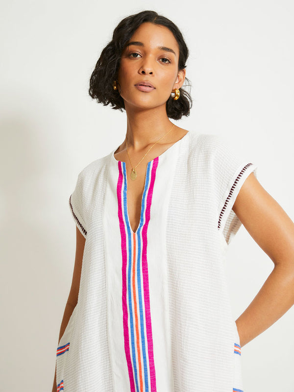 Close up on a Woman Standing Wearing lemlem Elina Caftan featuring stripe pattern of bright magenta, blue, and orange on s crispy white background. 