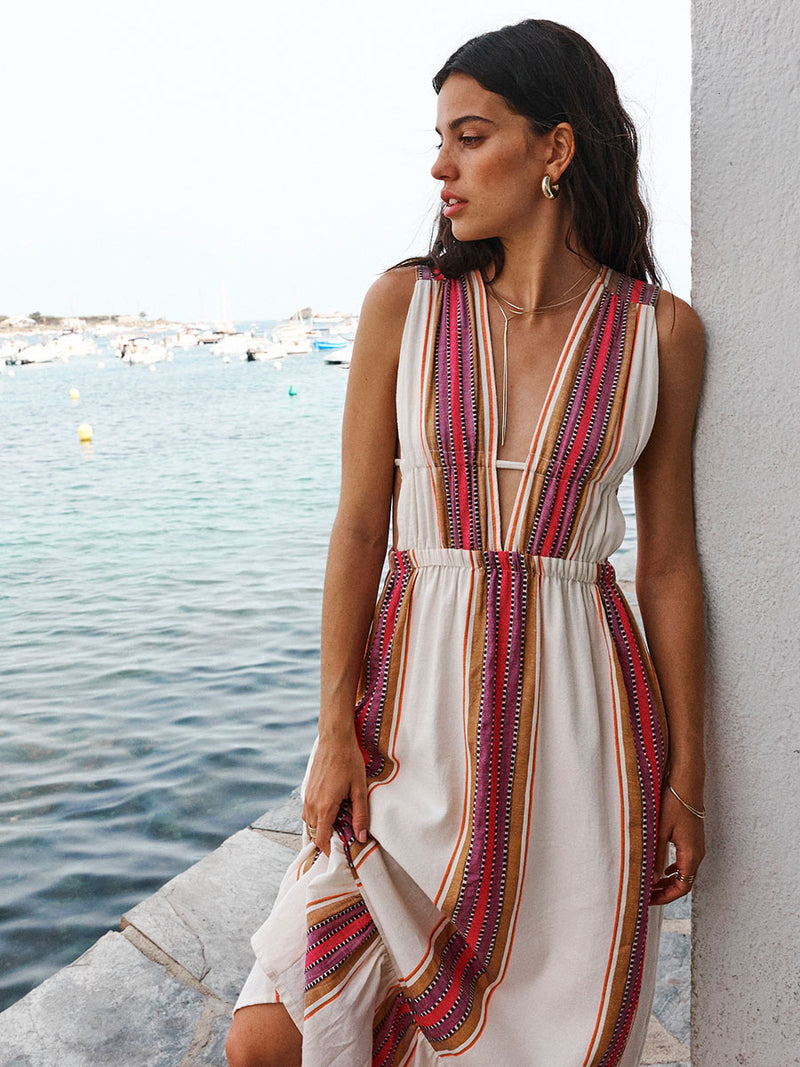 Woman standing against a white wall by the water wearing a long plunge neck dress in ivory with brown and berry graphic stripes. 