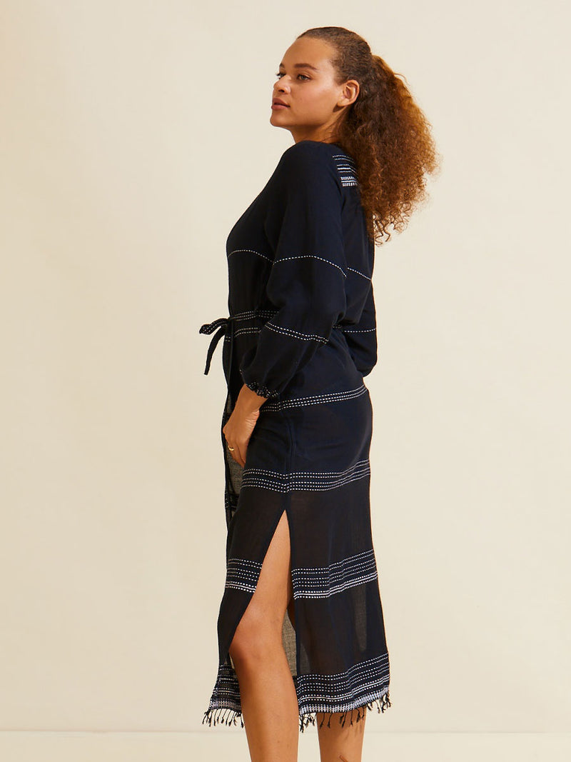 Side view of a Woman standing wearing the Leliti Long Robe in black with white stitching allover and matching waist tie.