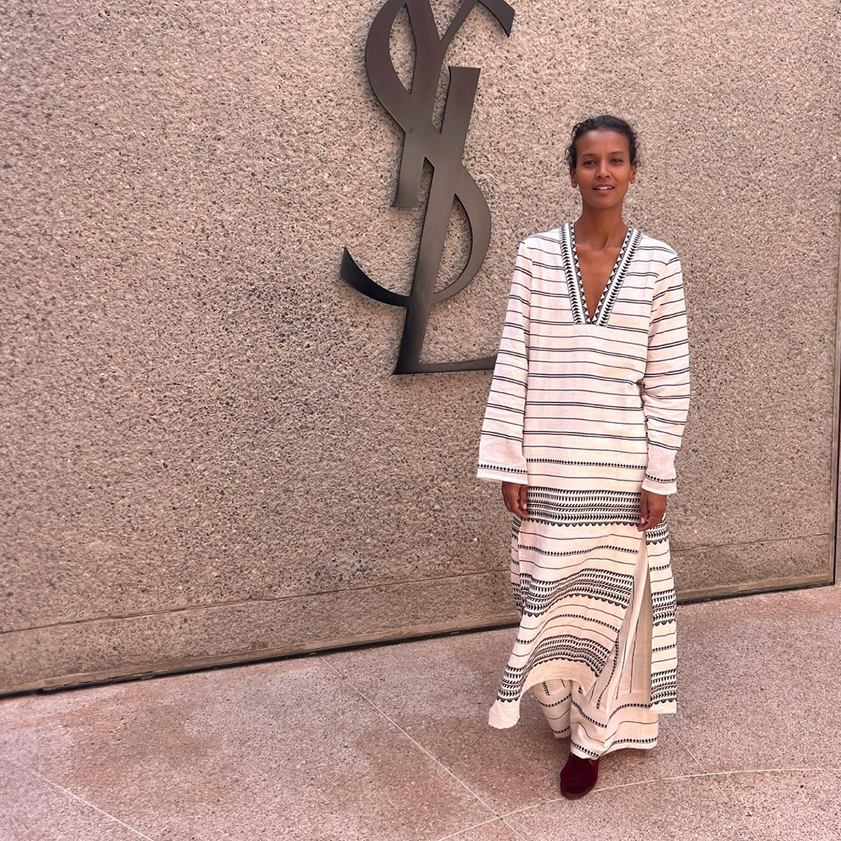 Liya Kebede wearing a long lemlem caftan in white with black graphic over matching wide leg lemlem pants in front the YSL house. 
