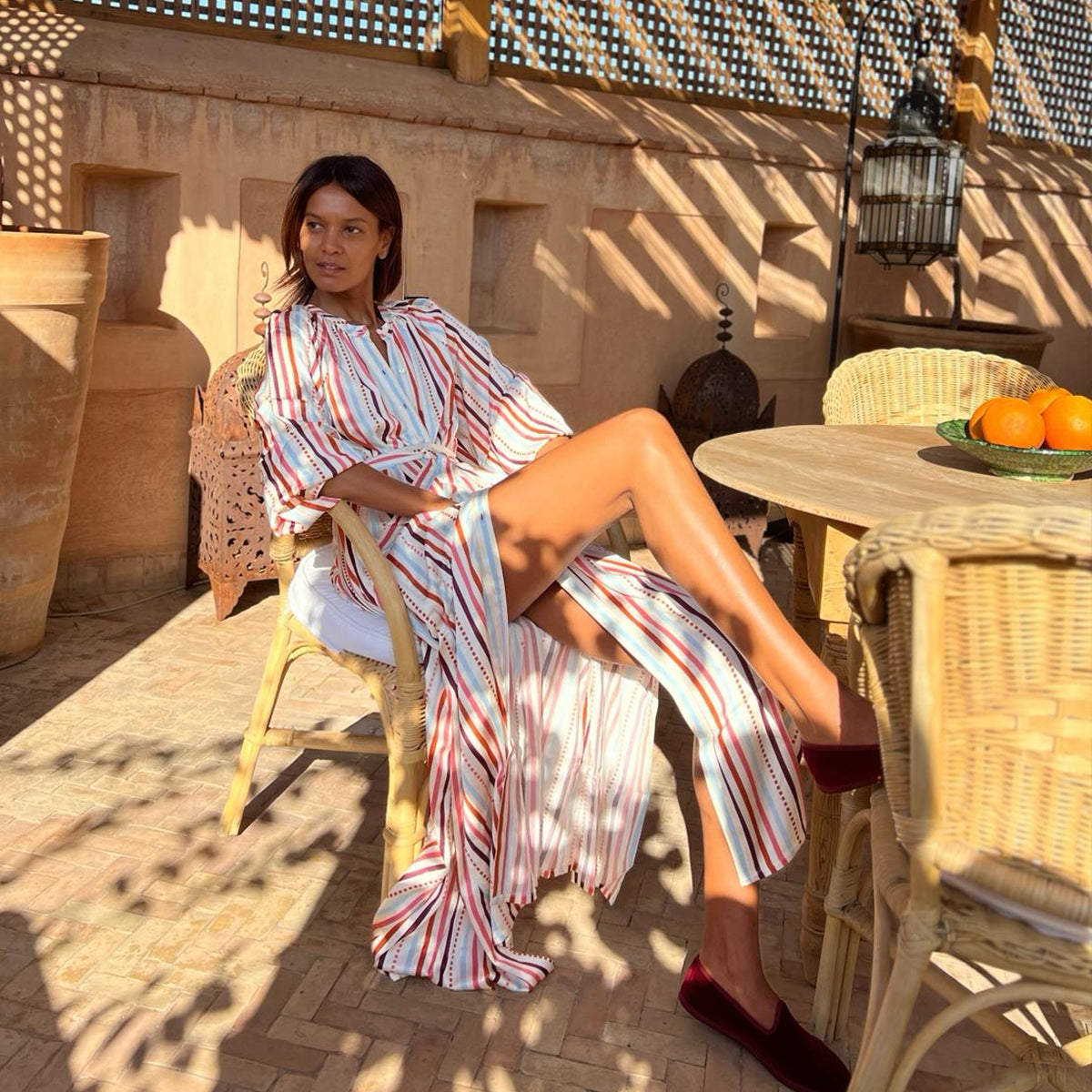 Liya Kebede lounging on a chiar with her legs crossed wearing a long white lemlem shirt dress in white with multi color stripes.