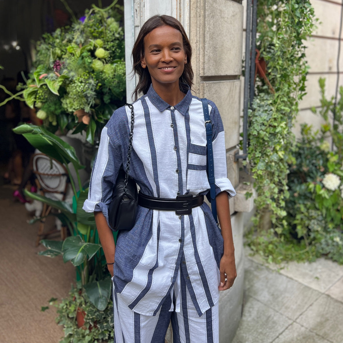 Liya Kebede standing in front of a wall covers in flowers wearing a lemlem blue and white oversized shirt and matching lemlem wide leg pants. 