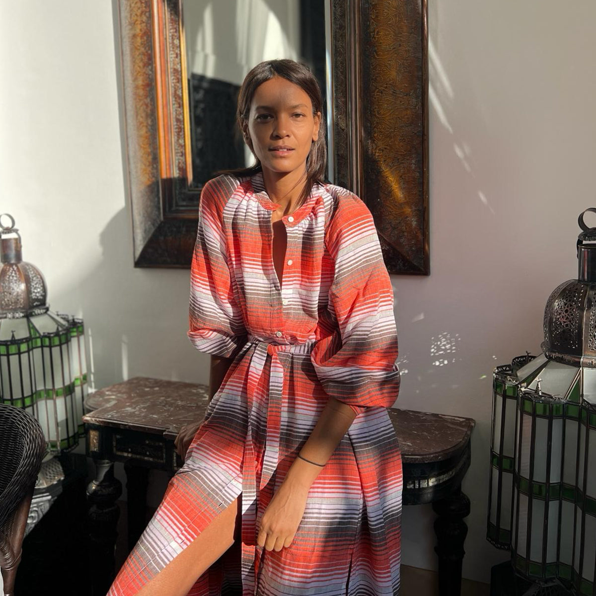 Liya Kebede sitting on a bench wearing a long lemlem shirt dress in red, white and brown small stripes.  