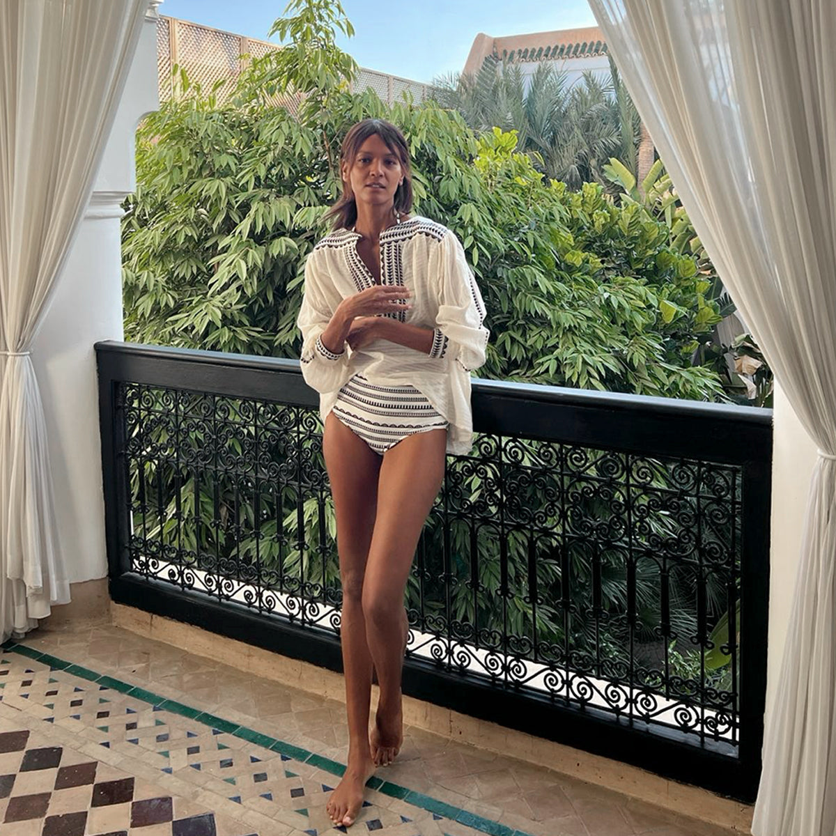 Liya Kebede standing on a balcony with lush trees in the background wearing a lemlem blouse in white with graphic black stripes and matching lemlem high waist bikini bottoms. 