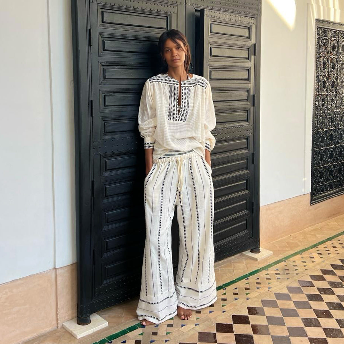 Liya Kebede standing in front a black door wearing a lemlem blouse in white with graphic black lines and matching lemlem wide leg pants. 