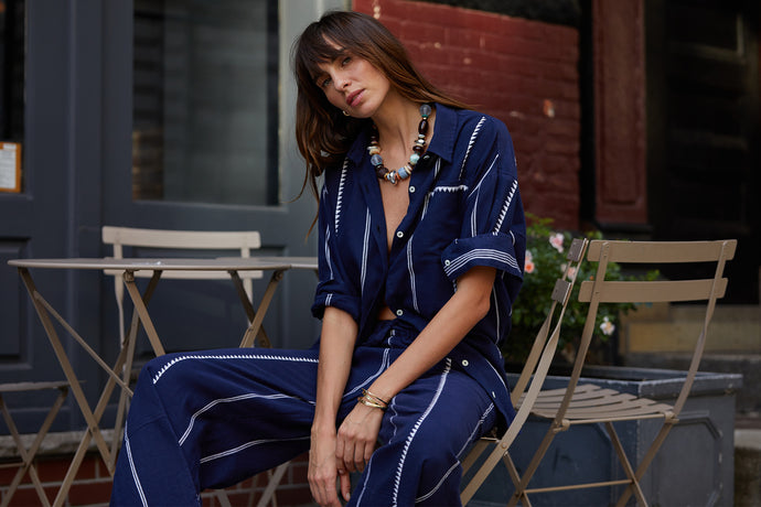 Woman sitting at a table wearing a lemlem handwoven blue bottom down shirt and matching handwoven pants.