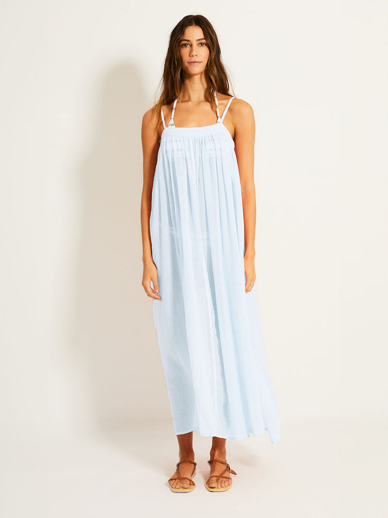 Woman standing wearing lemlem handwoven EDA Slip Dress featuring pale blue color and light sheer fabric