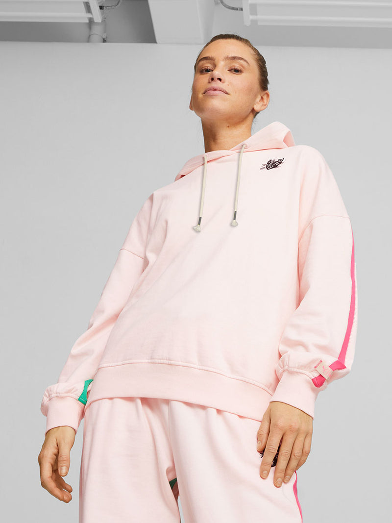Woman standing wearing Puma x lemlem fleece oversized hoodie and joggers in Ghost Pepper Color featuring color pop side stripes in pink and green colors and handsketched puma cat logo