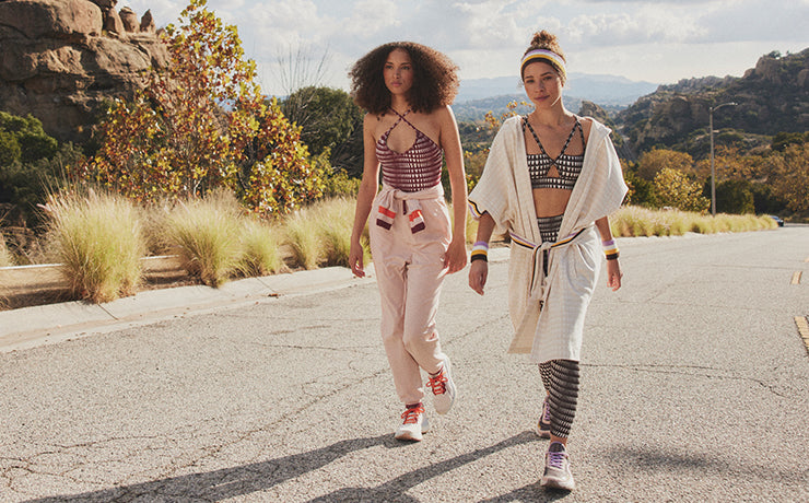 Two Woman walking on an empty road wearing the Puma x lemlem collaboration. Two women sitting on a rock wearing the Puma x lemlem 2024 collaboration. One is wearing the Puma x lemlem low impact bra and matchin Puma x lemlem legging in brown with a the white puma x lemlem anorak and Puma x lemlem PWR NITRO™ SQD Women's Training Shoes. The other is wearing the Puma x lemlem jumpsuit with burgundy Puma x lemlem leotard and Puma x lemlem ForeverRun NITRO™ Women's Running Shoes.