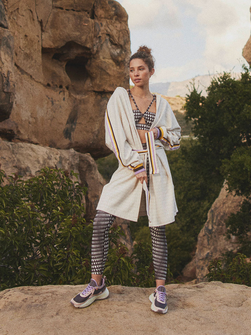 Woman Standing Wearing Puma x lemlem Anorak in Warm White Color, featuring lemlem triangle pattern and color stripe details in violet, dark chocolate, white and yellow colors and puma x lemlem leggings and bra in  dark chocolate colors 