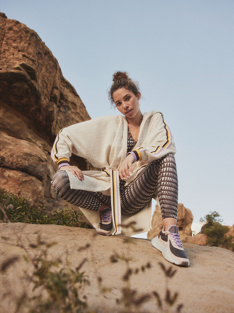 Woman Squatting Wearing Puma x lemlem Anorak in Warm White Color, featuring lemlem triangle pattern and color stripe details in violet, dark chocolate, white and yellow colors and puma x lemlem leggings and bra in dark chocolate colors