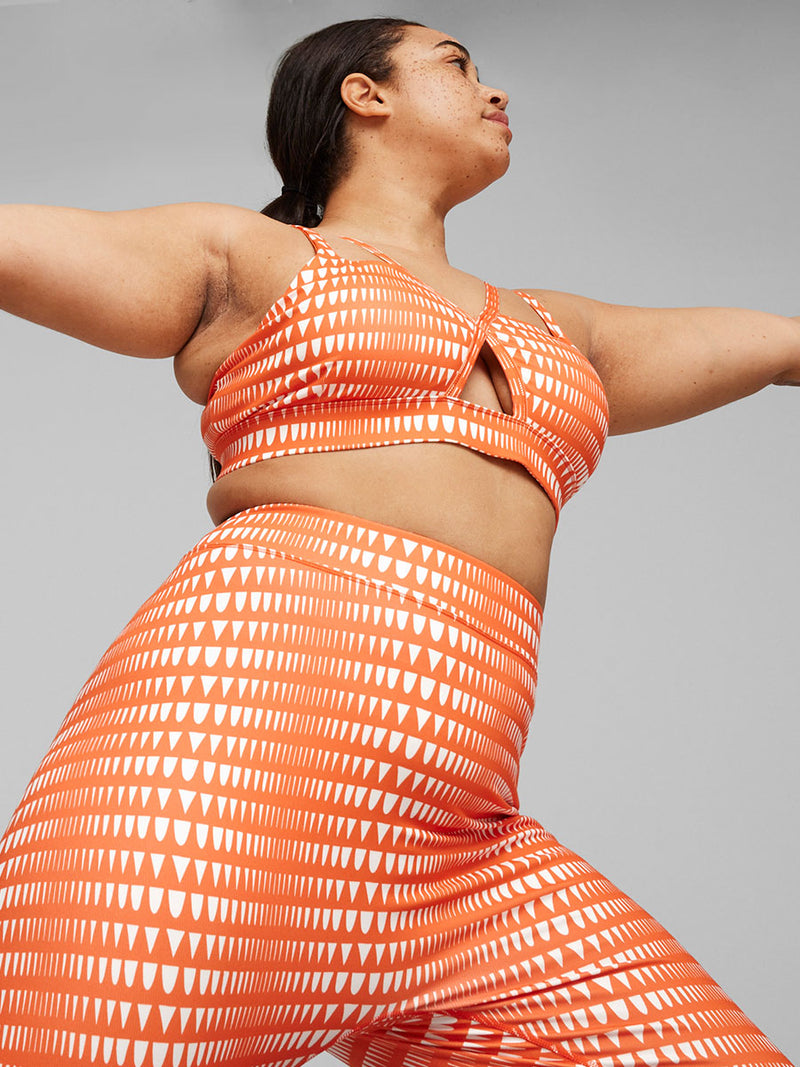 Side View of Woman Standing Wearing  Puma x lemlem Low Impact Bra in Cayenne Pepper Color Featuring lemlem Triangle Pattern in White Color and matching high waist leggings