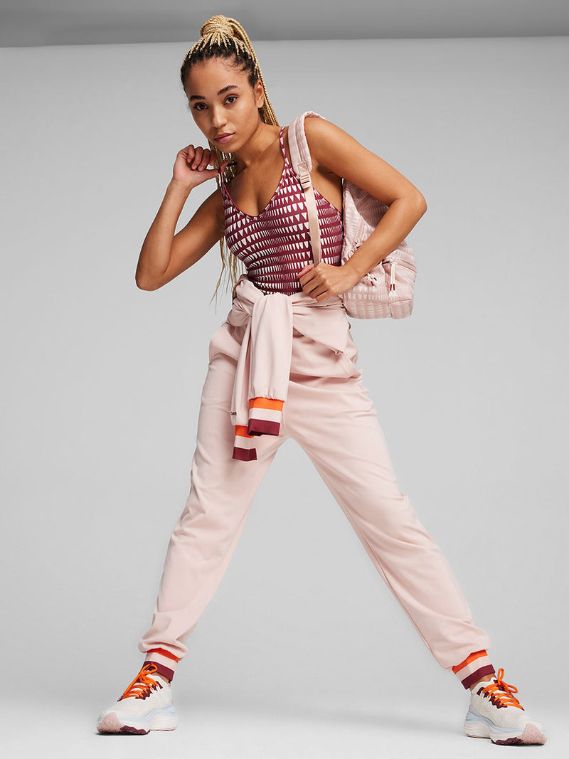 Woman Standing Wearing Puma x lemlem Jumpsuit featuring Rose Quartz Color, color block details in orange and red colors on collar, arf cuffs and ankle cuffs, puma x lemlem leotard, running shoes and pink backpack