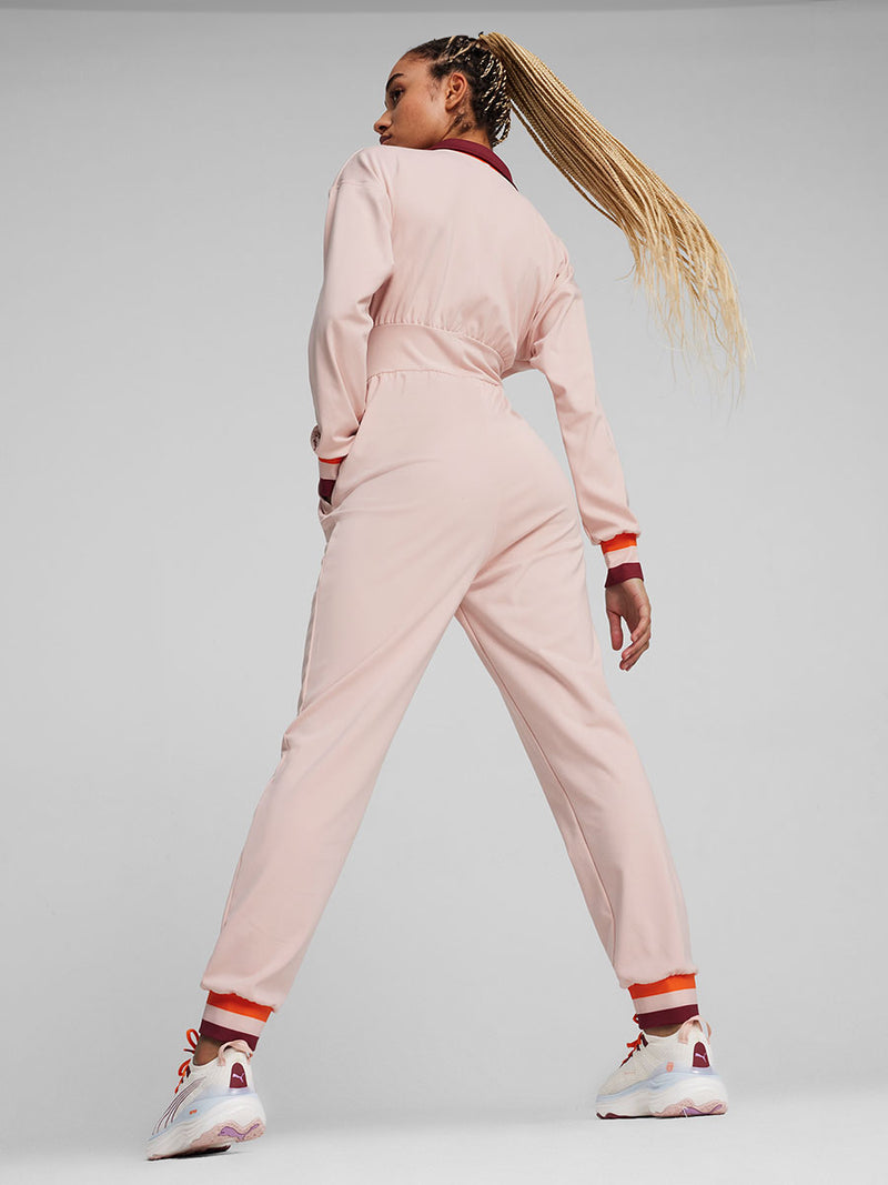 Back View of a Woman Standing Wearing Puma x lemlem Jumpsuit featuring Rose Quartz Color, color block details in orange and red colors on collar, arf cuffs and ankle cuffs and puma x lemlem nitro running shoes